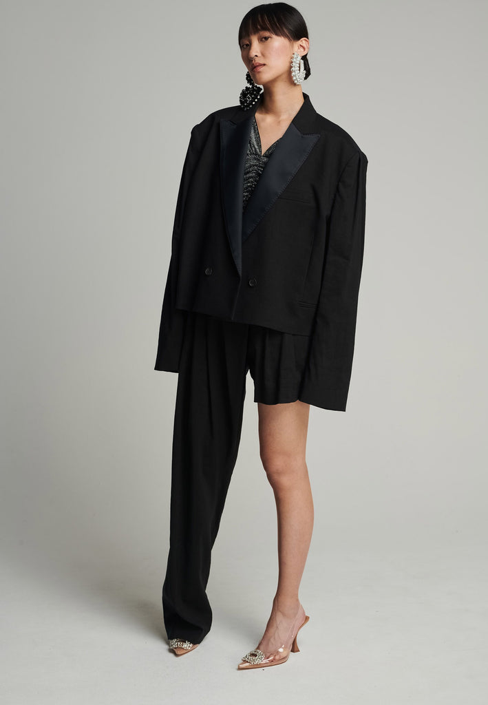 Wide-shouldered short blazer in black. Features shoulder-pads, pockets, and exaggerated long sleeves. Fits oversize