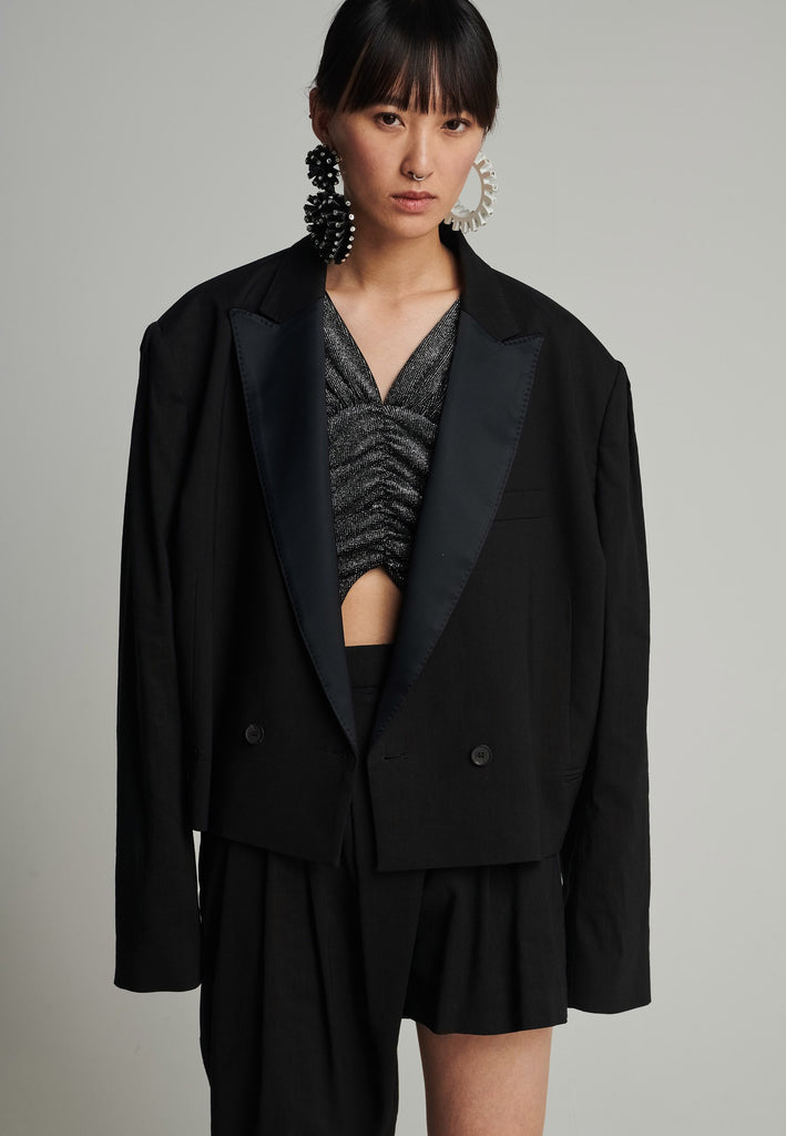 Wide-shouldered short blazer in black. Features shoulder-pads, pockets, and exaggerated long sleeves. Fits oversize